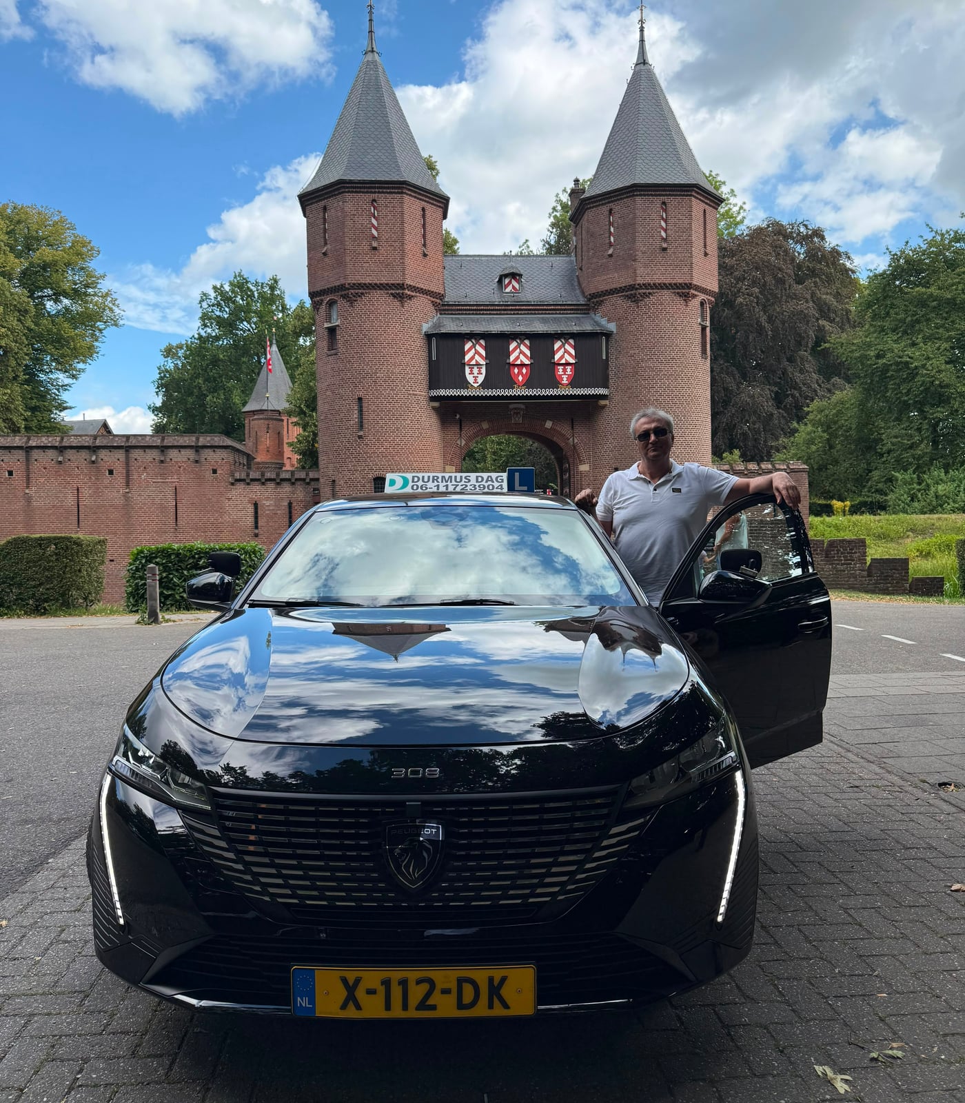 Driving instructor Durmus Dag beside the open door of the black Peugeot lesson car, in front of the gatehouse of De Haar Castle